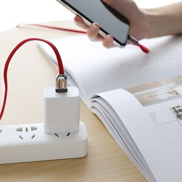  کابل تبدیل USB به لایتنینگ هوکو مدل U74 به طول 1.2 متر که به یک شارژر دیواری متصل شده و گوشی هوشمند را در کنار یک دفترچه شارژ می‌کند. 